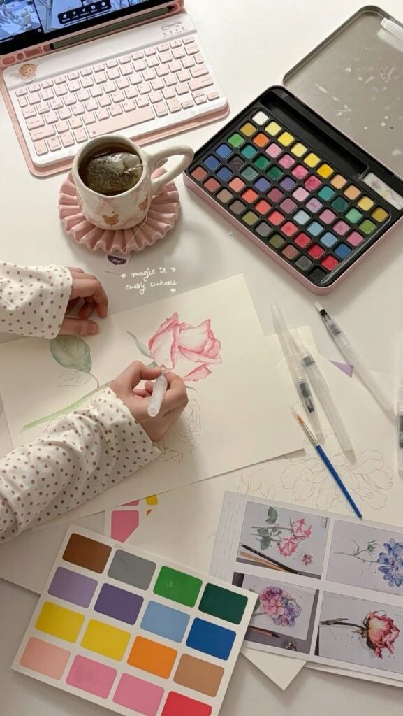 Artist sketching rose with pink watercolor on desk with color palette, brushes, tea, and laptop nearby.