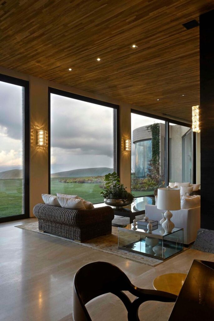 Modern living room with large windows, wicker furniture, and scenic views of a green landscape under cloudy skies.