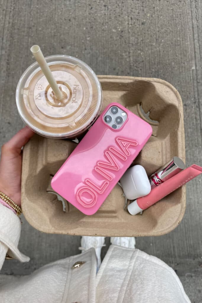 Person holds iced coffee and tray with pink phone case, AirPods, and lip gloss on a street background.