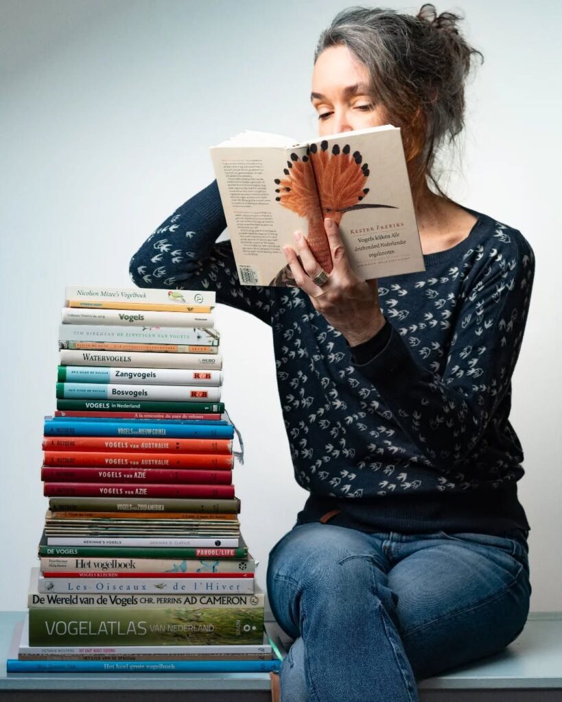Woman reading a book on birds, seated next to a tall stack of ornithology books.
