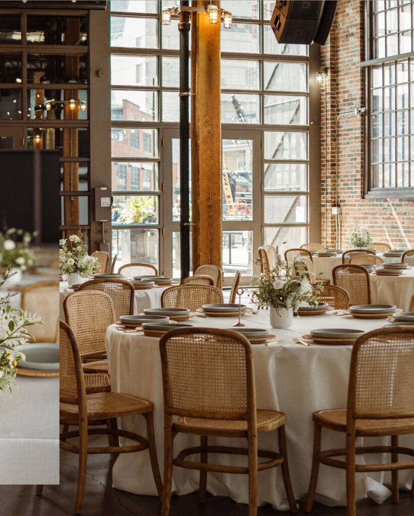 Elegant event space with round tables, white linens, wicker chairs, floral centerpieces, and large windows.