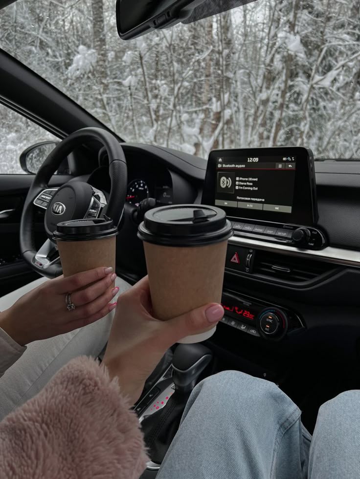 ba9d834a-b686-448b-96f0-4703a27550f9 Two people enjoy takeaway coffee inside a car, parked in a snowy forest setting, with music playing on the dashboard. | Sky Rye Design Two people enjoy takeaway coffee inside a car, parked in a snowy forest setting, with music playing on the dashboard.