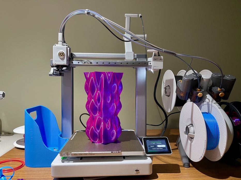 bambua1-cover3s-1 3D printer crafting a vibrant purple object with spooled filament beside it, on a wooden desk. | Sky Rye Design 3D printer crafting a vibrant purple object with spooled filament beside it, on a wooden desk.