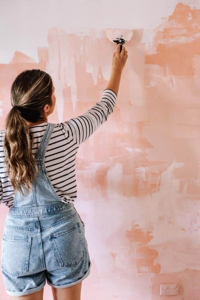Woman painting wall with pink brush strokes, wearing striped shirt and denim overalls. Home DIY project.