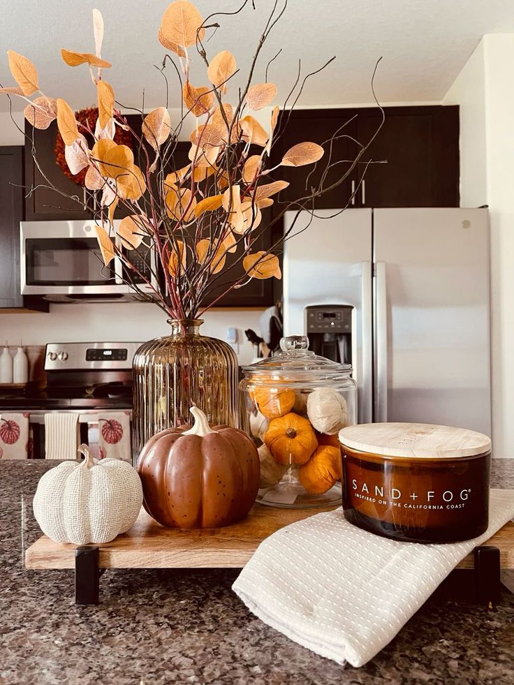 Autumn kitchen decor with vase, dried leaves, pumpkins, candle, and towel on a granite countertop. Cozy fall ambiance.