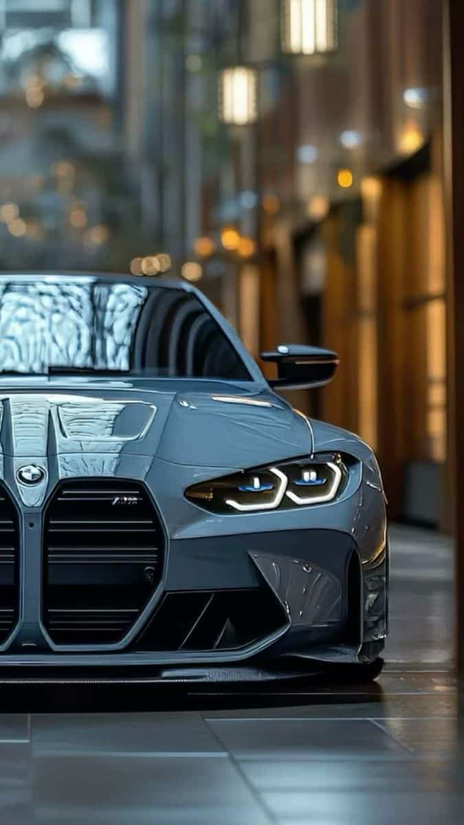 Close-up of a sleek gray BMW M4 sports car with illuminated headlights in an urban setting.