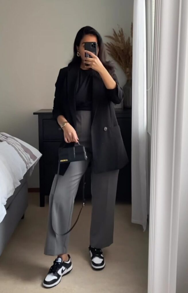Woman in black blazer and gray pants takes mirror selfie, accessorized with small bag and sneakers in stylish room.