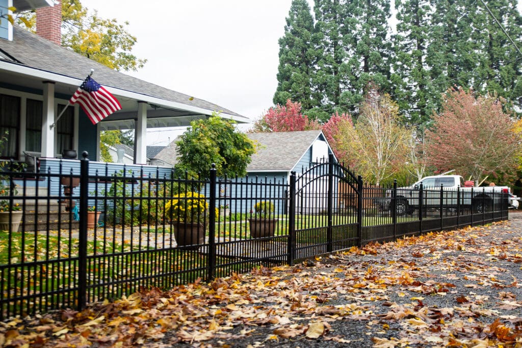 best-fencing-company-near-me Blue house in autumn with an American flag and black iron fence, surrounded by colorful trees and fallen leaves. | Sky Rye Design Blue house in autumn with an American flag and black iron fence, surrounded by colorful trees and fallen leaves.
