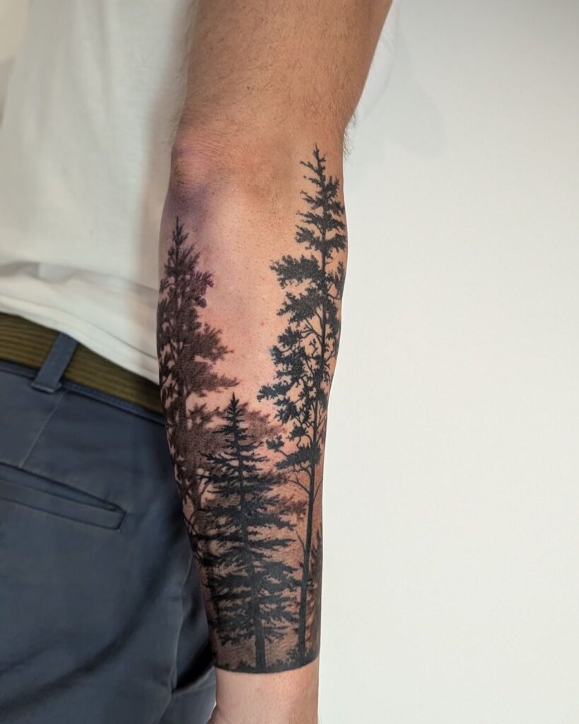 Forearm tattoo featuring detailed pine trees in black ink against a sunset-inspired background.