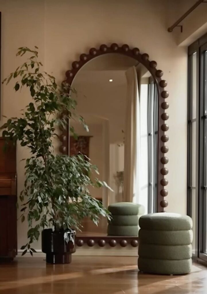 Stylish interior with large mirror, lush potted plant, and cozy pouf near a window.