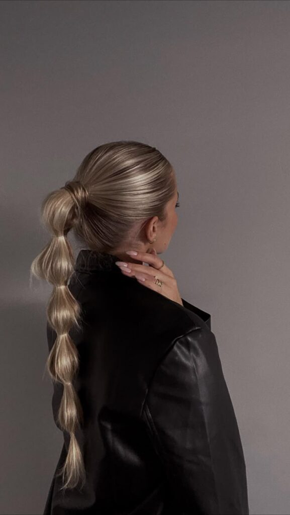 bubble ponytail 🫧 using kenraprofessional Elegant ponytail hairstyle with blonde waves, woman in black jacket, side view against a neutral background. | Sky Rye Design Elegant ponytail hairstyle with blonde waves, woman in black jacket, side view against a neutral background.