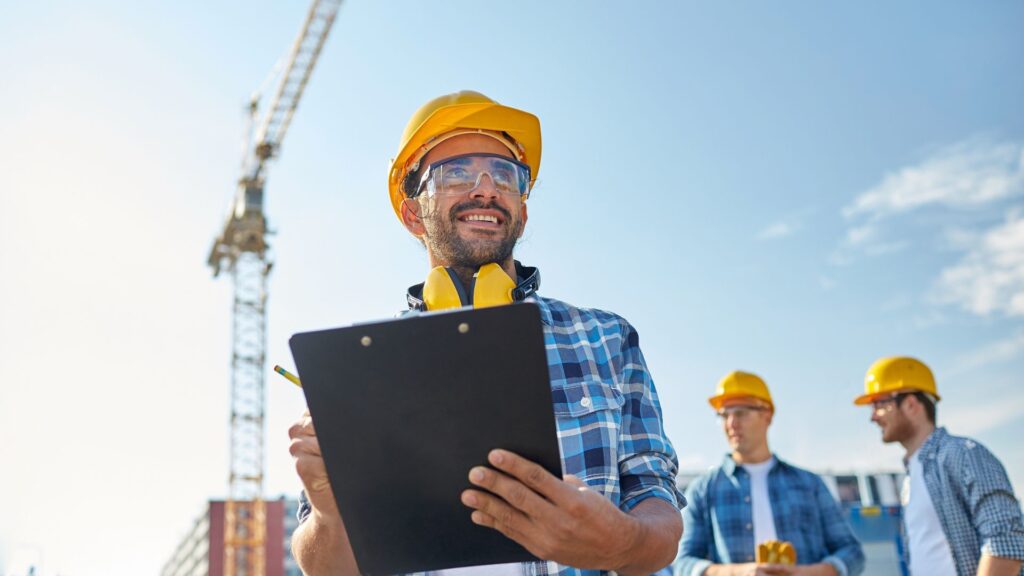 builder-5 Construction worker in a hard hat smiling with clipboard outdoors, crane and colleagues in background. | Sky Rye Design Construction worker in a hard hat smiling with clipboard outdoors, crane and colleagues in background.