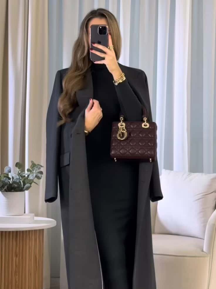 Elegant woman in a black outfit with a long coat, holding a stylish dark handbag, taking a mirror selfie.