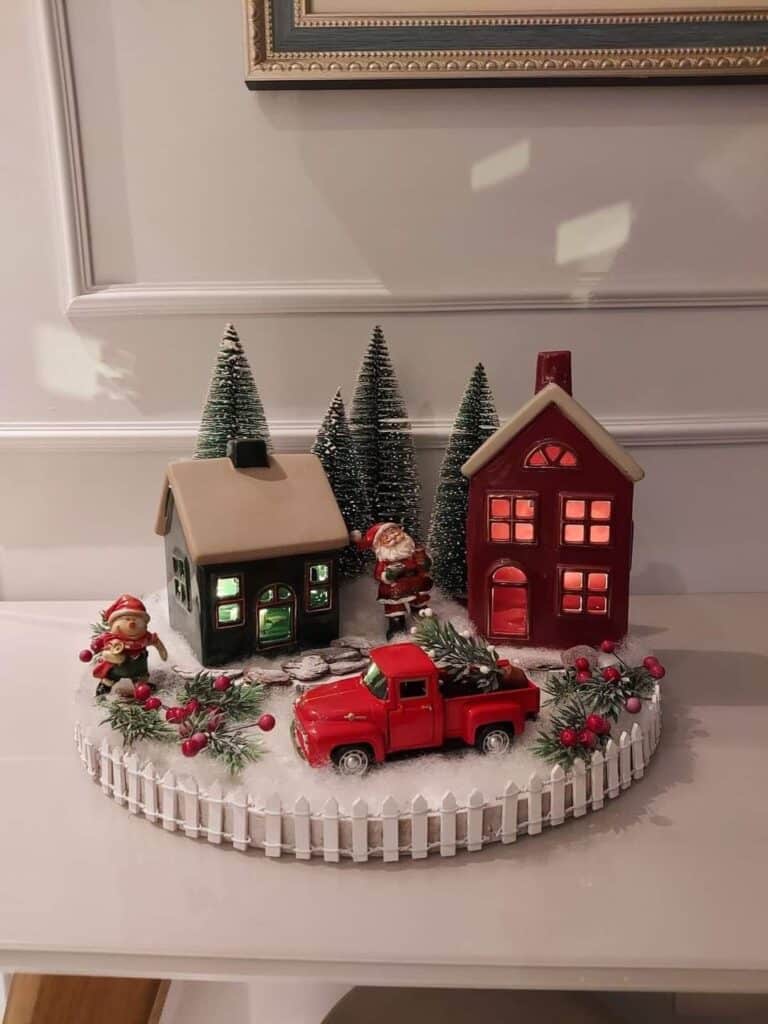 c16cf5ef58c53f1b3eb503ff72ee751c Festive Christmas village scene with Santa, houses, red truck, and snowy trees behind a white picket fence. | Sky Rye Design Festive Christmas village scene with Santa, houses, red truck, and snowy trees behind a white picket fence.