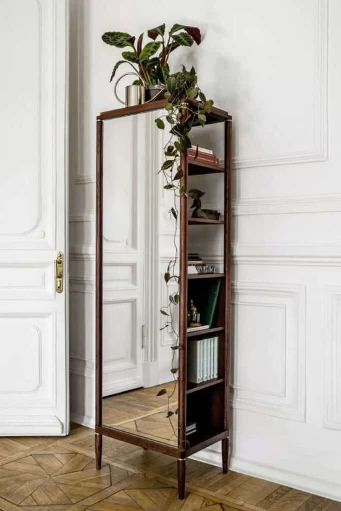 Tall mirror with shelves holding books and plants, set against an elegant white wall for a chic interior look.