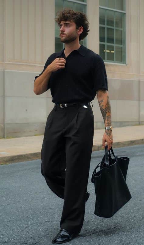 Man in stylish black outfit walks confidently with a tote bag on city street.