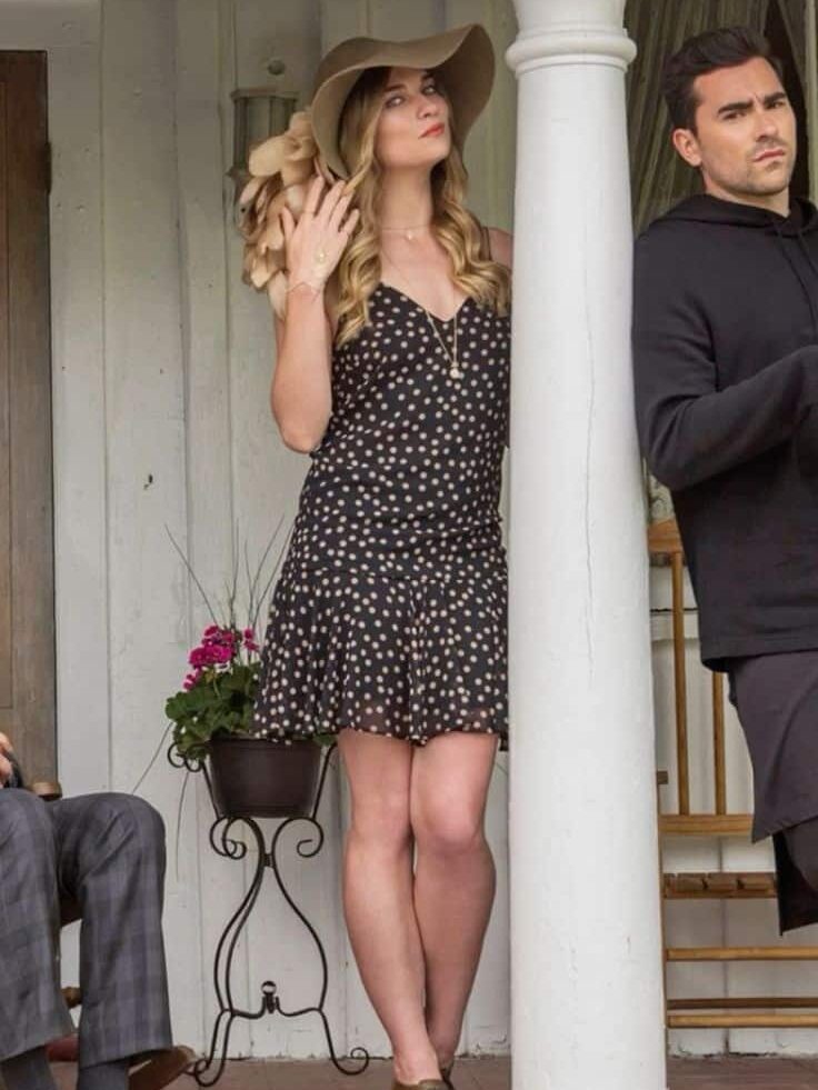 Woman in polka dot dress and hat stands on porch, next to a man in black outfit, both near a white column.