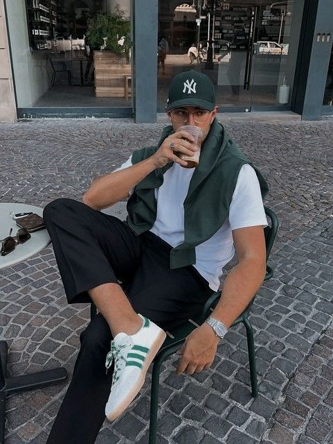 Casually dressed man in a hat enjoys a drink at an outdoor café, wearing stylish sneakers and black pants.