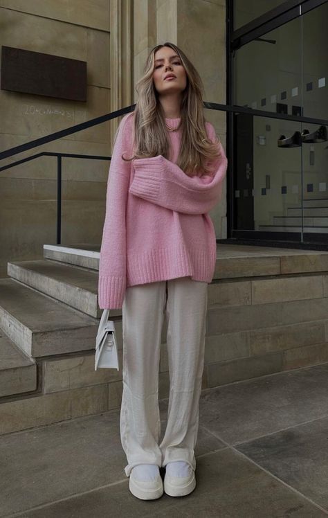 Woman in pink sweater and white pants stands outside on steps, holding a white bag. Fashionable and casual look.