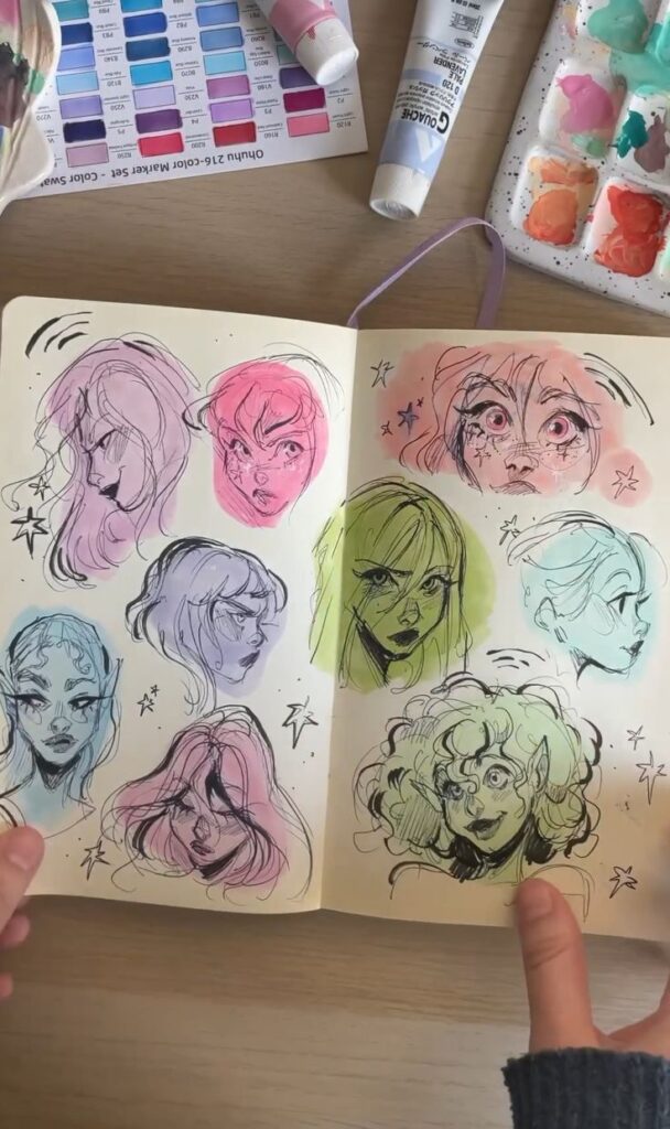 Sketchbook page with colorful character sketches, watercolor palette, and paint tubes on a wooden desk.
