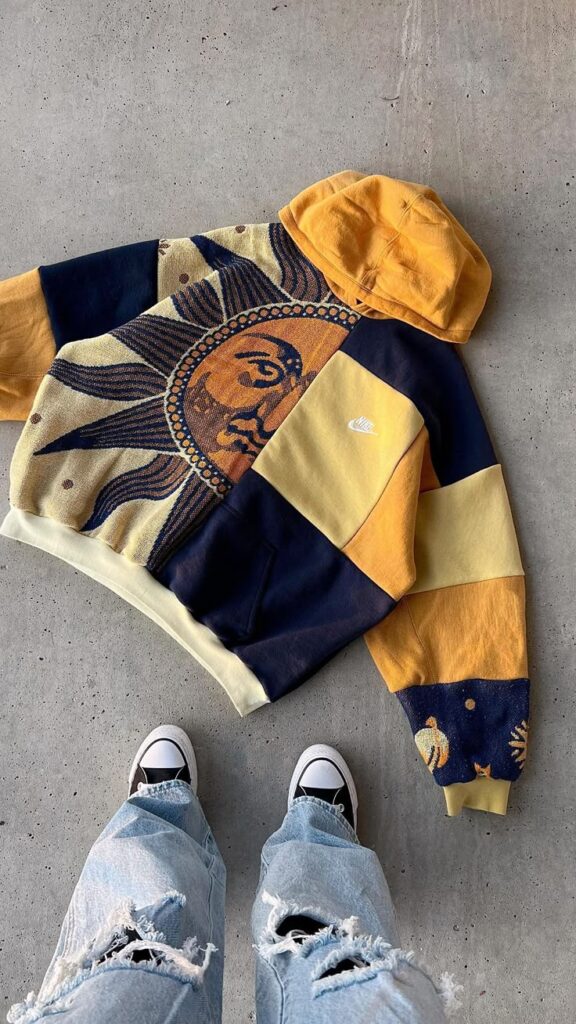 Colorful sun design hoodie with patchwork detail laid out on the ground with ripped jeans and sneakers visible.
