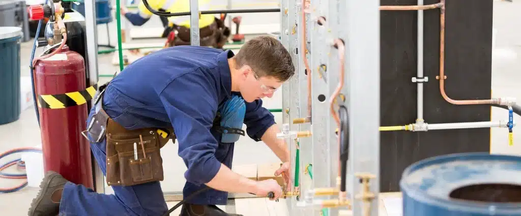 construction-plumbing Technician in blue uniform inspecting plumbing pipes indoors, wearing safety glasses. | Sky Rye Design Technician in blue uniform inspecting plumbing pipes indoors, wearing safety glasses.