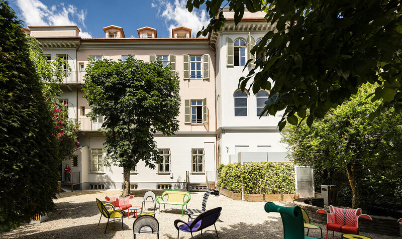 cover-video-sede-torino-quintino Sunny courtyard with colorful chairs and greenery outside a historic building. | Sky Rye Design Sunny courtyard with colorful chairs and greenery outside a historic building.