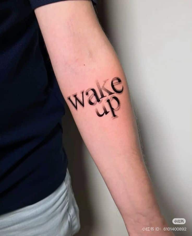 Tattoo on inner forearm reading 'wake up' in black font.