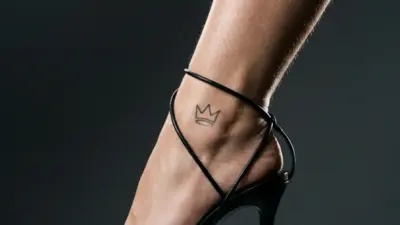 Close-up of woman's foot in black strappy high-heeled sandal on marble pedestal with small crown ankle tattoo