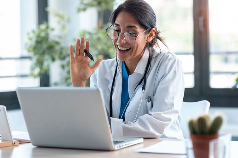 customer-support-for-virtual-medical-assistant Doctor having a video call on a laptop, smiling and waving, in a bright office. | Sky Rye Design Doctor having a video call on a laptop, smiling and waving, in a bright office.