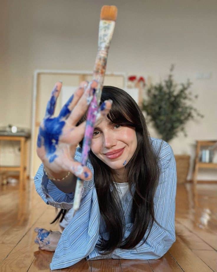 Smiling woman lying on floor, holding paintbrush with blue paint on hand, indoor art activity.