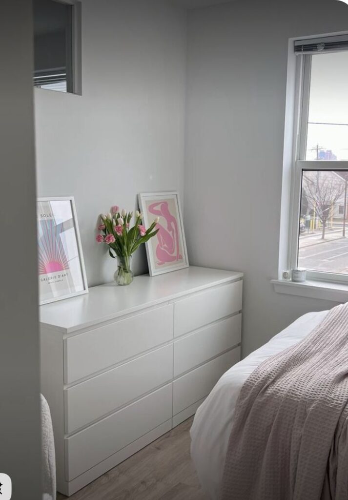 d2db35c1-19e4-4323-b1e3-97d0242bb5de Minimalist bedroom with white dresser, pink tulips, and window view. Contemporary art and cozy bed setting. | Sky Rye Design Minimalist bedroom with white dresser, pink tulips, and window view. Contemporary art and cozy bed setting.