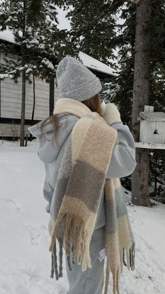 Person in winter clothes with scarf and beanie, standing in snowy forest near white cabin.