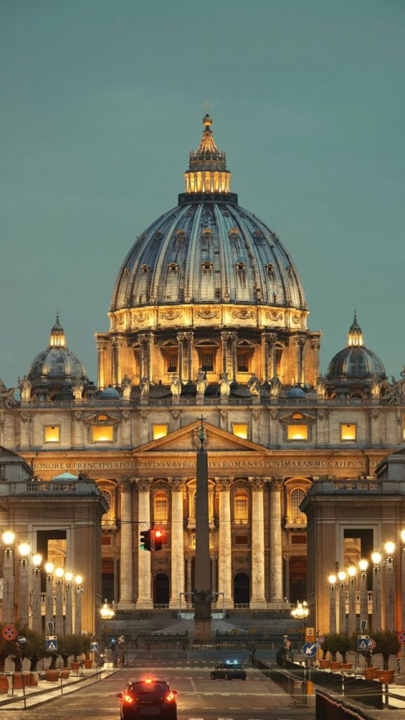 St. Peter's Basilica illuminated at dusk, Vatican City, with street lights and cars in the foreground.