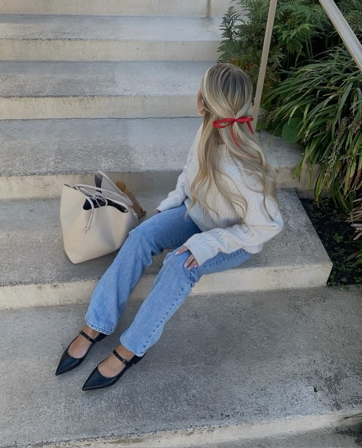 d6b6a184-cb98-4369-beda-886d85608ce5 Woman with red hair ribbon in sweater and jeans sits on steps by greenery, holding a beige tote bag. | Sky Rye Design Woman with red hair ribbon in sweater and jeans sits on steps by greenery, holding a beige tote bag.