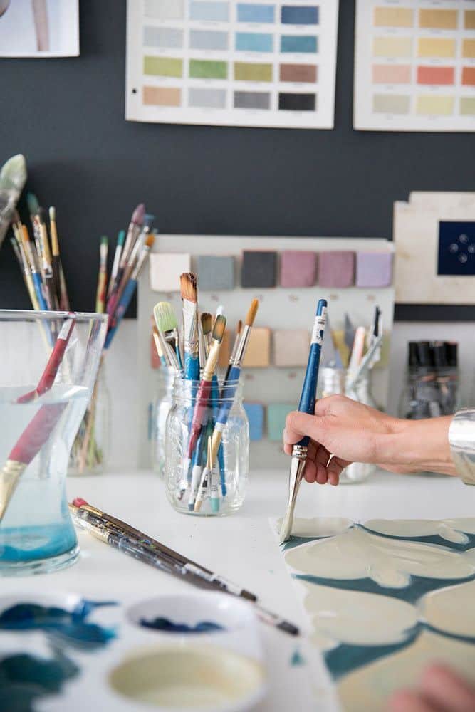 Artist painting with brushes and color samples on a table; creative workspace filled with art supplies.