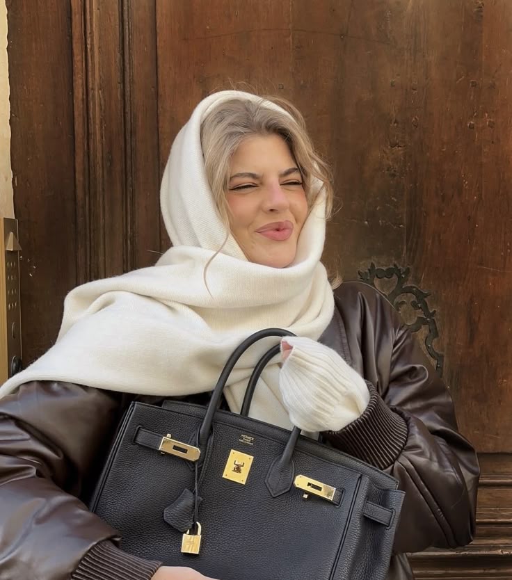Woman in winter attire smiles holding a luxury handbag against a wooden backdrop.