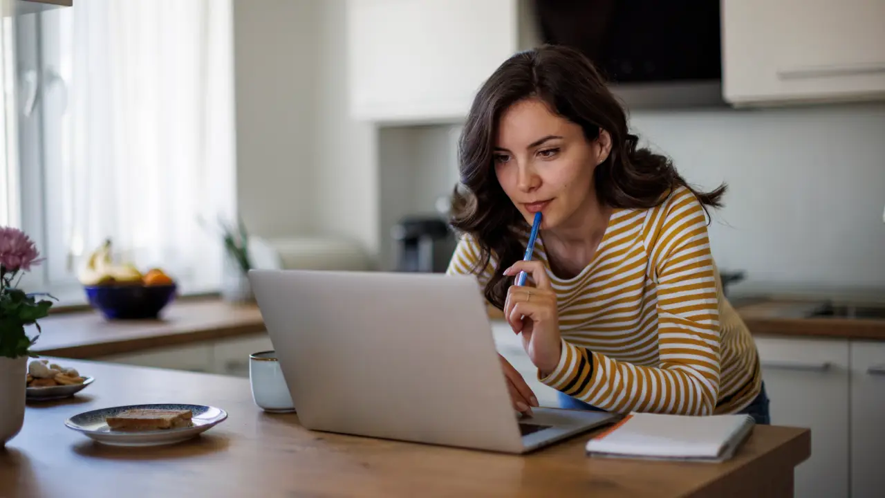 debt-relief-guide Woman in striped shirt working on a laptop at a kitchen table, thoughtful expression with a pen in hand. | Sky Rye Design Woman in striped shirt working on a laptop at a kitchen table, thoughtful expression with a pen in hand.