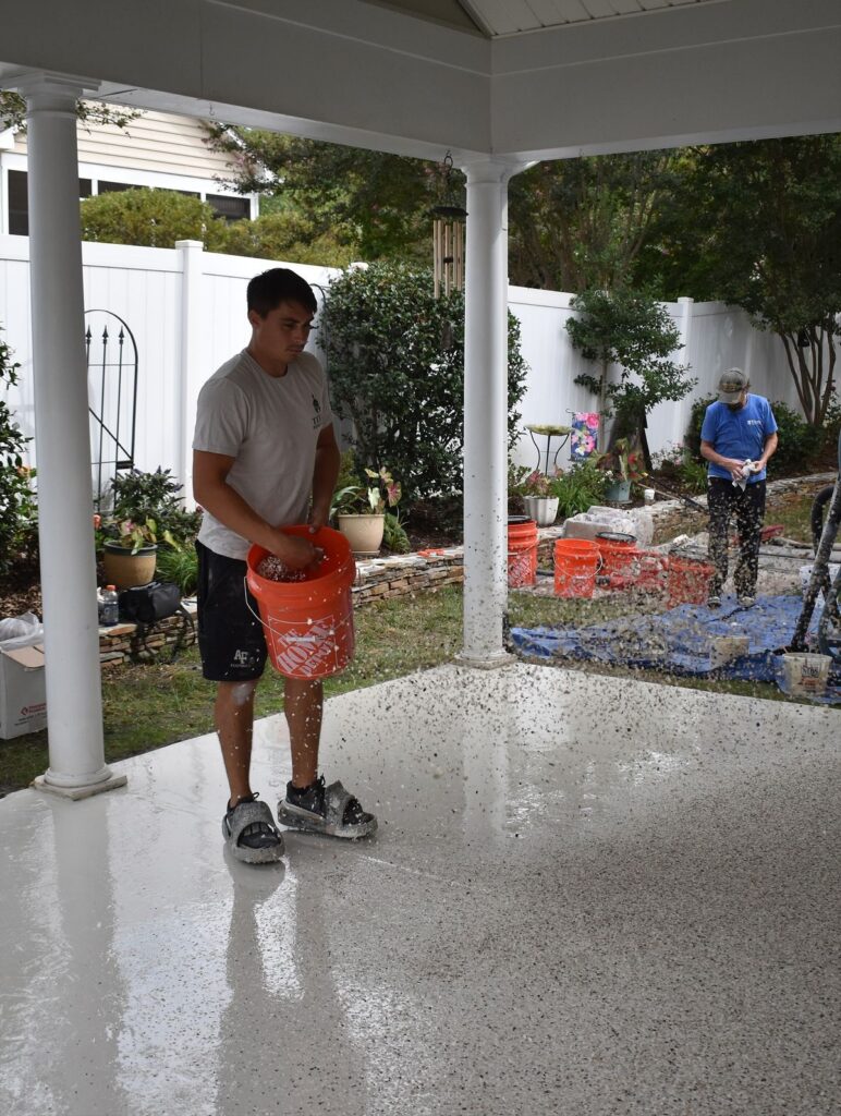 Person scattering paint chips on epoxy garage floor for anti-slip surface under porch.