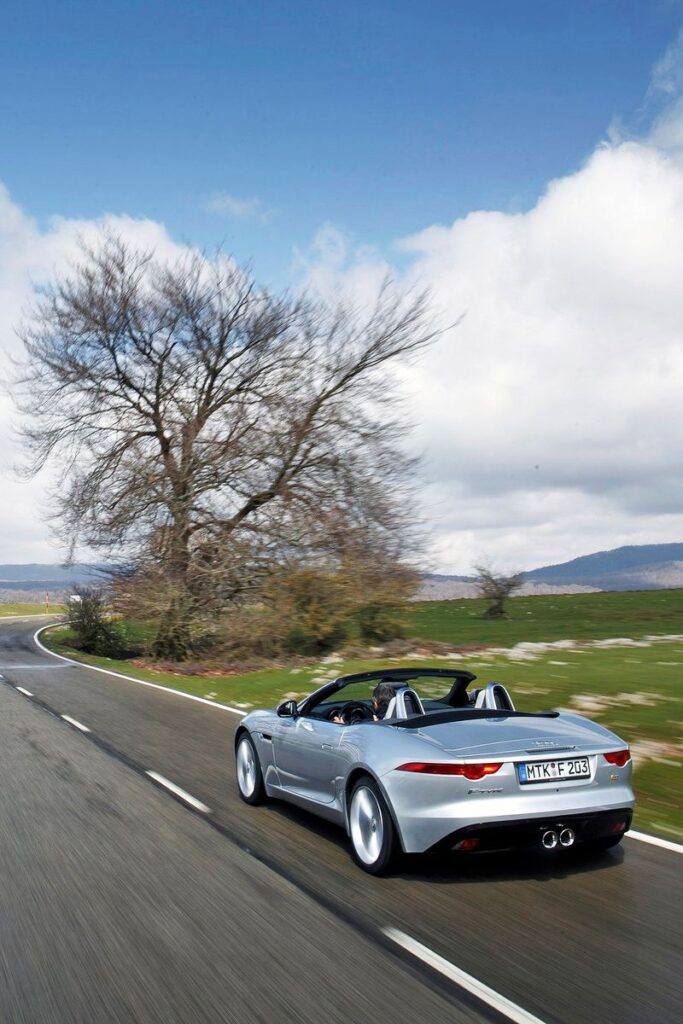 Sports car driving on scenic road with trees and mountains under a blue sky.