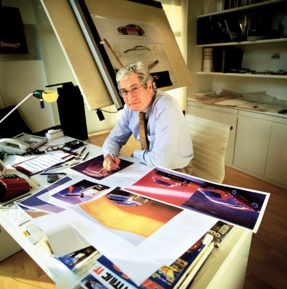 Designer at work in an office, reviewing car designs and sketches on a cluttered desk.