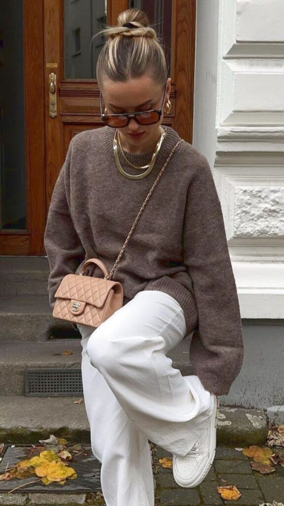 Fashionable woman in neutral tones with sunglasses, wearing a brown sweater, white pants, and gold jewelry.