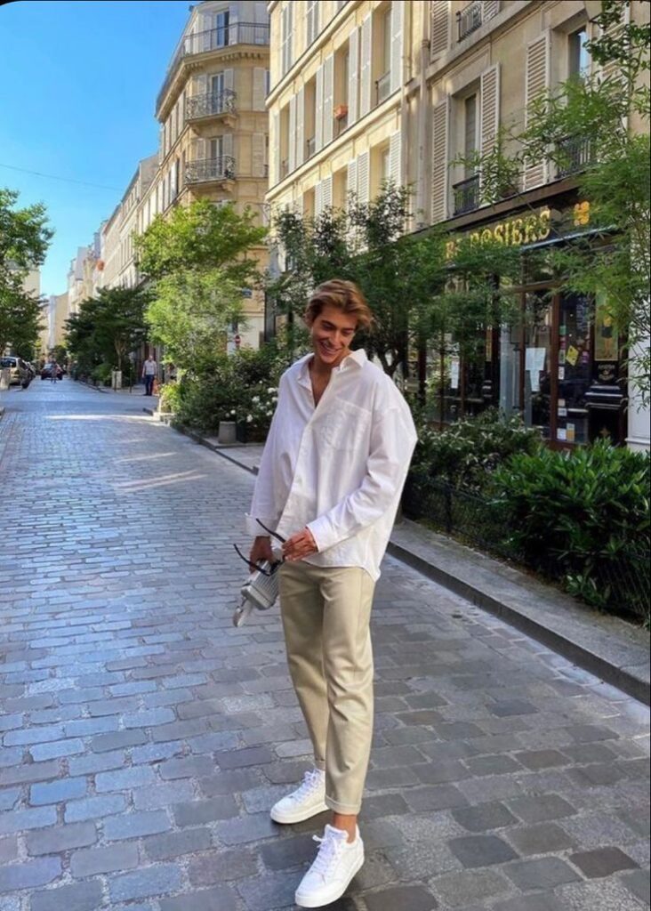 Person smiling while walking on a European street in casual attire, holding a vintage camera, surrounded by buildings and greenery.