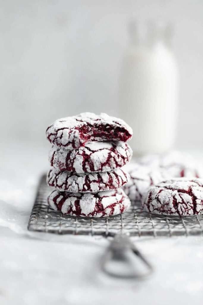 Stack of red velvet crinkle cookies dusted with powdered sugar on a cooling rack with a bottle of milk in the background.