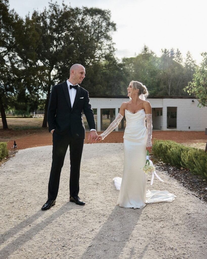 Bride and groom holding hands outdoors, smiling, dressed in formal attire on a sunny day.
