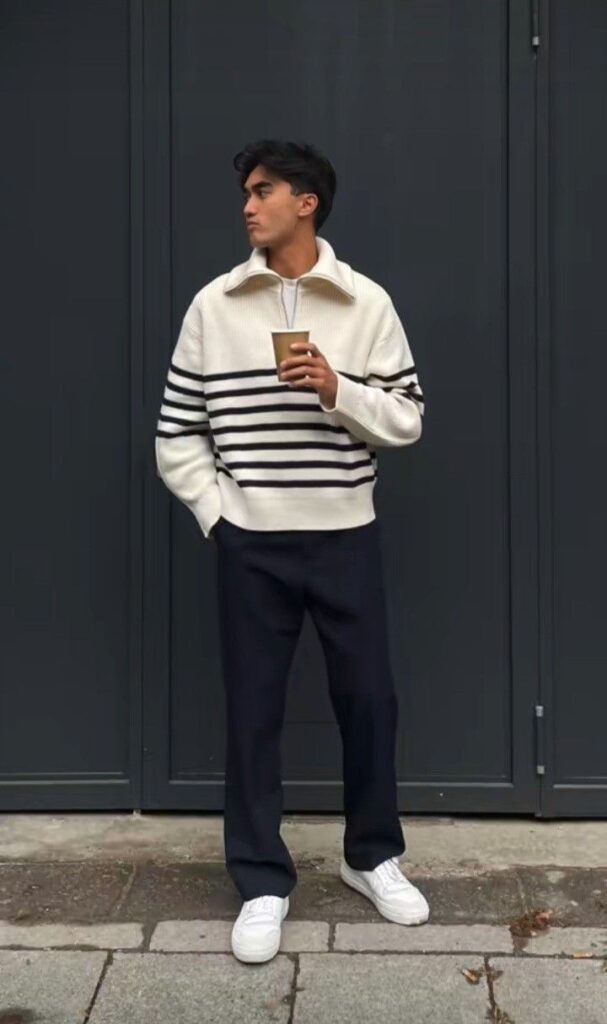 f0181607-e3dd-4ac6-b9d1-403944e3b33b Man wearing a striped sweater and holding a coffee cup stands against a dark wall. Casual street fashion. | Sky Rye Design Man wearing a striped sweater and holding a coffee cup stands against a dark wall. Casual street fashion.