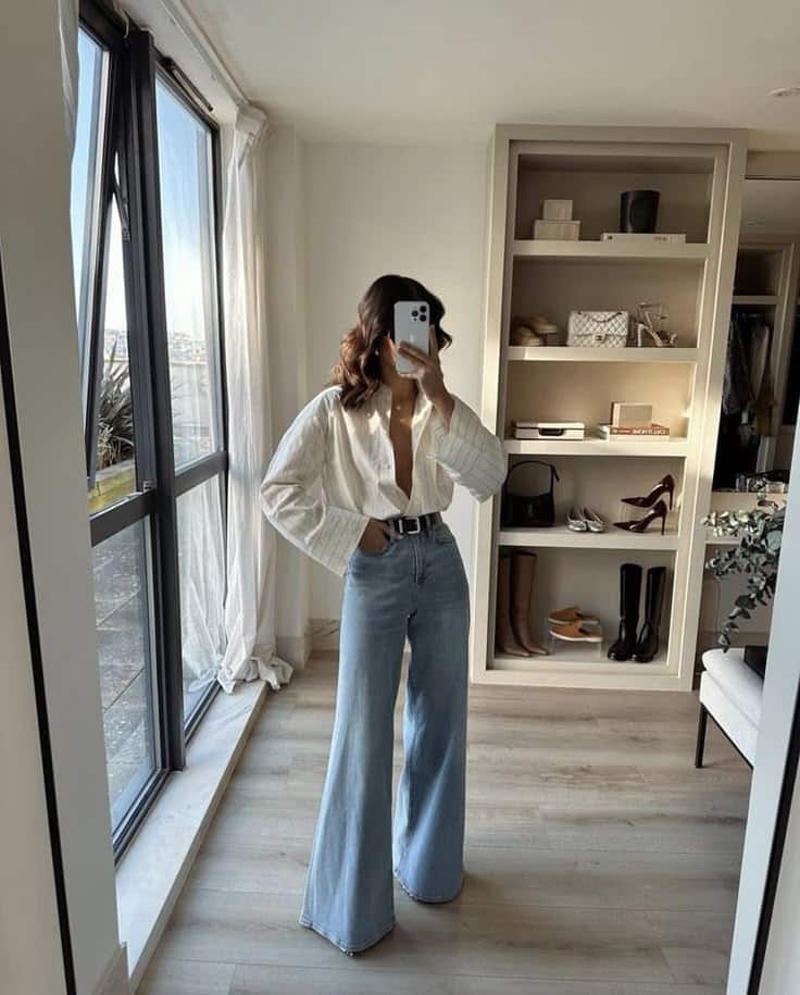 Fashionable woman in flared jeans and white top takes a mirror selfie in chic, sunlit room with open closet shelving.