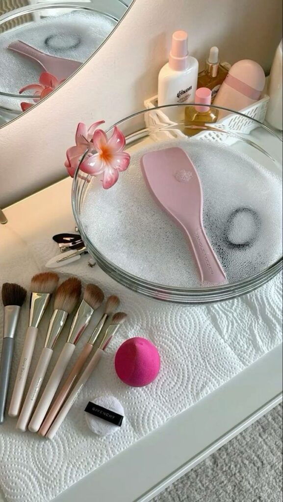 f54ccd70-8b62-4a87-87ba-8572950aae85 Makeup brushes and tools being cleaned in soapy water, with products and accessories on a vanity table. | Sky Rye Design Makeup brushes and tools being cleaned in soapy water, with products and accessories on a vanity table.