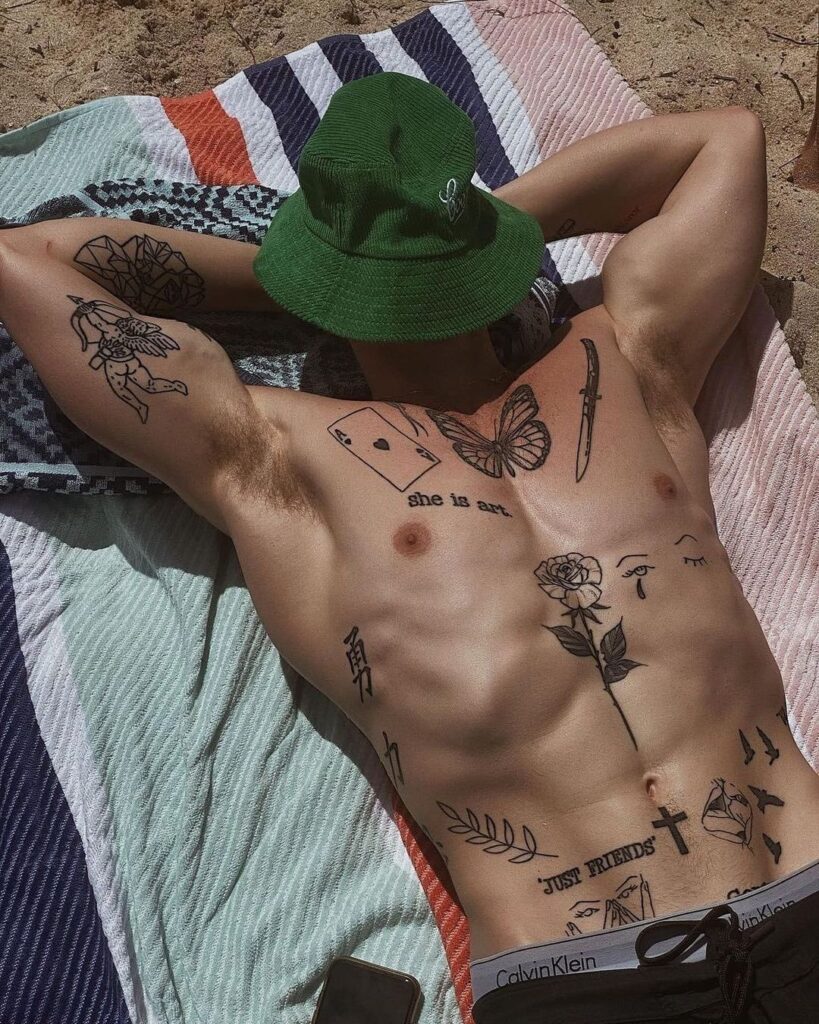 f57afe0fcb96d640f9a0e80b9b3452c0 Man with tattoos sunbathing on beach, wearing green hat, lying on striped blanket. | Sky Rye Design Man with tattoos sunbathing on beach, wearing green hat, lying on striped blanket.