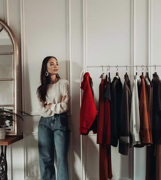 f65b9783-c497-4c95-a2a4-549620d35847 Woman in casual attire standing by a rack of assorted clothing against a stylish room backdrop. | Sky Rye Design Woman in casual attire standing by a rack of assorted clothing against a stylish room backdrop.